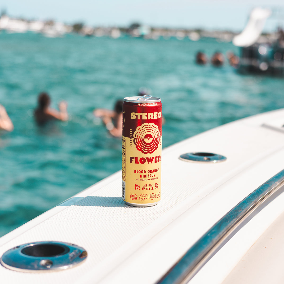 Main - A can of Stereo Flower Blood Orange Hibiscus alcoholic beverage stands on the white deck of a boat with turquoise water and swimmers blurred in the background. - 2