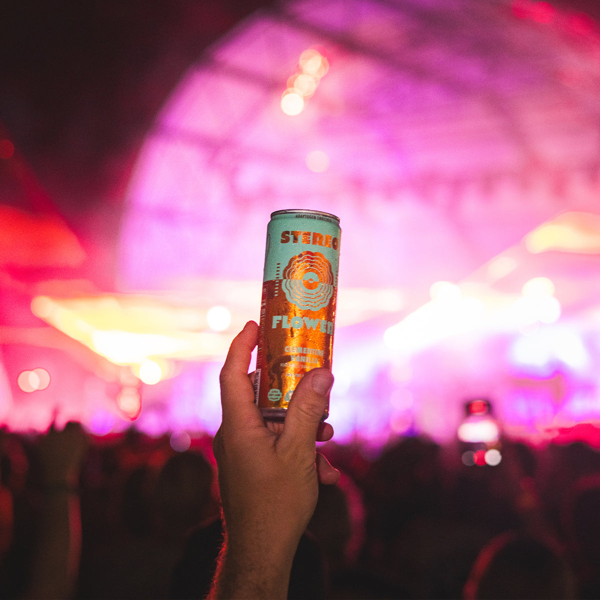 A hand holds up a can of Stereo Flower beverage at a concert with a blurred stage in the background.