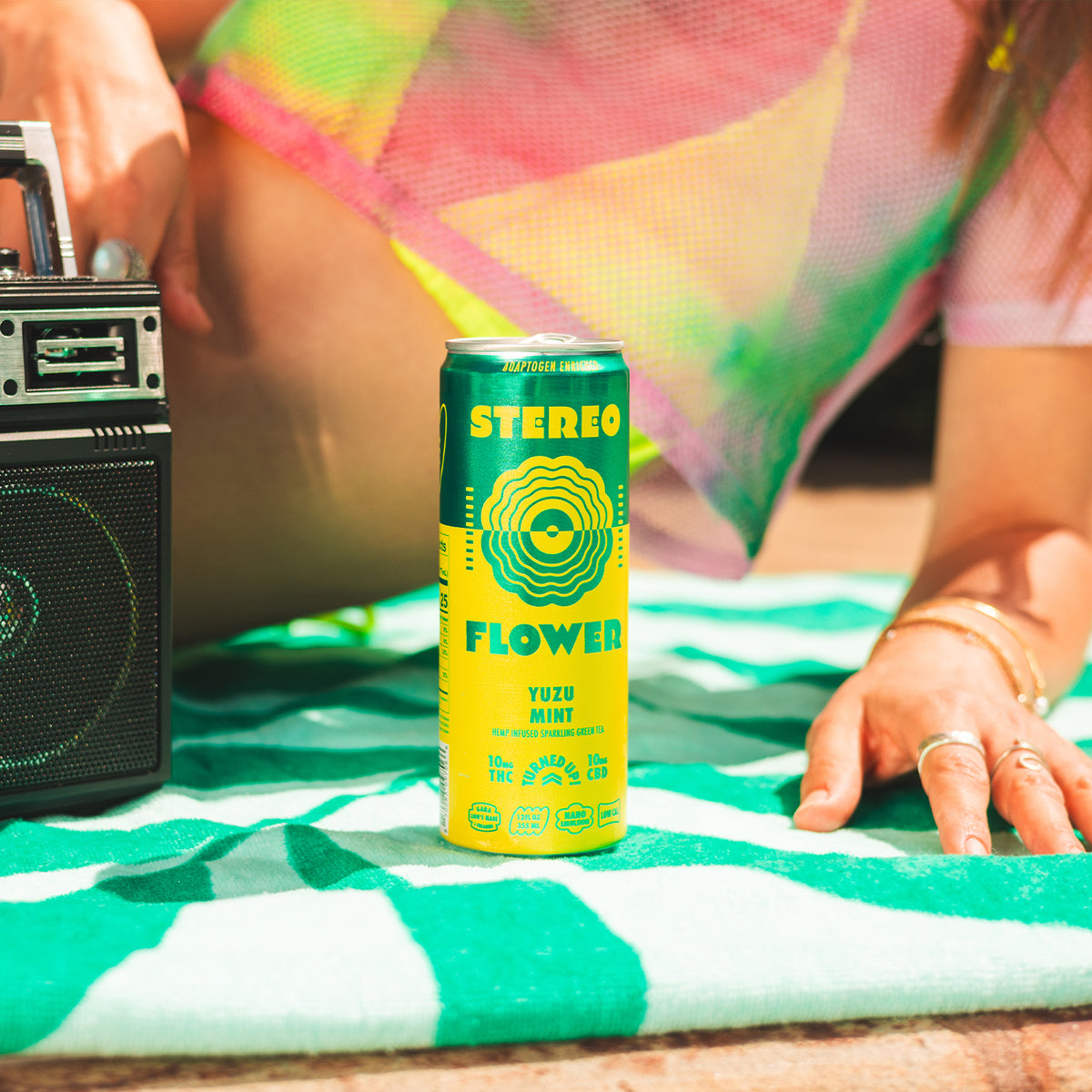 Main - A yellow and green can of Stereo Flower Yuzu Mint beverage stands on a green and white striped blanket next to a vintage radio, with a person's hands and colorful clothing visible in the background. - 2