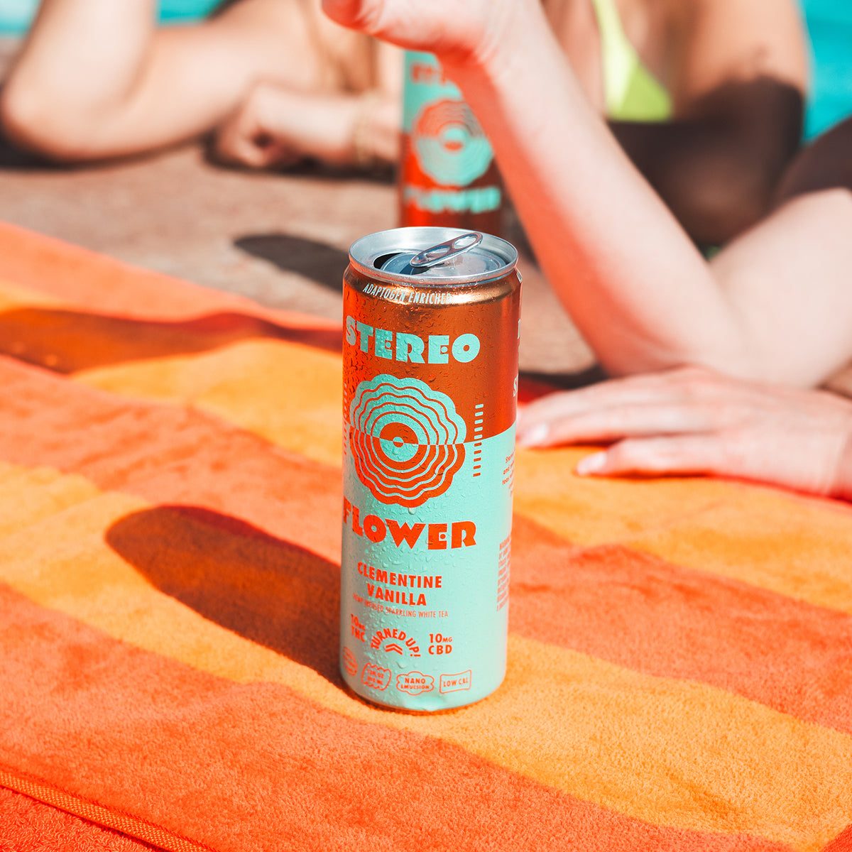 Main - A can of Stereo Flower Clementine Vanilla soda sits on an orange striped beach towel, with a person lying in the background near a pool. - 2