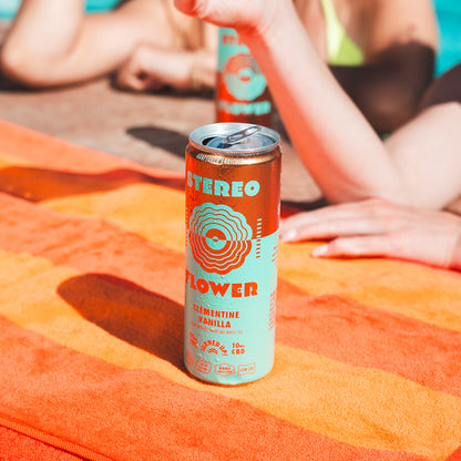 Thumbnail - A can of Stereo Flower Clementine Vanilla soda sits on an orange striped beach towel, with a person lying in the background near a pool. - 2