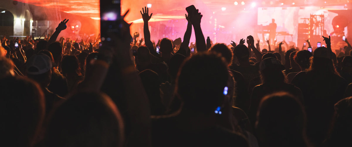 A crowd of concert-goers with raised hands in front of a brightly lit stage at a live music performance, with orange and pink stage lighting and silhouetted figures.